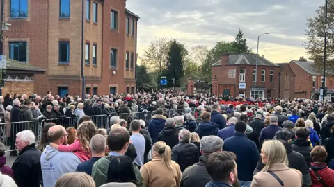 A crowd of hundreds of people line the streets of a town centre with houses and buildings in the background.