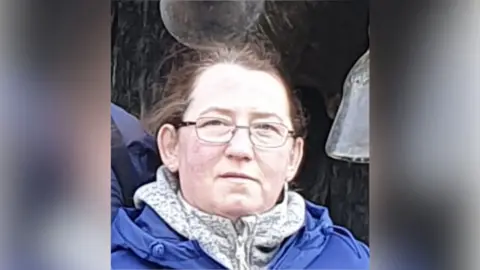 Northamptonshire Police Marta Bednarczyk with medium-length brown hair and thin-rimmed glasses. She is wearing a blue raincoat over a white and grey top