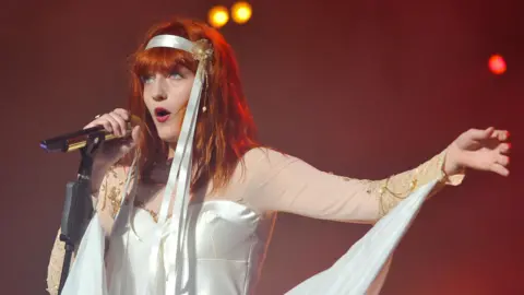 Andy Sheppard/Redferns The lead singer from Florence and the Machine is performing onstage. She is singing into a microphone. She has long red hair tied with a white ribbon and is wearing a long sleeveless white dress. 