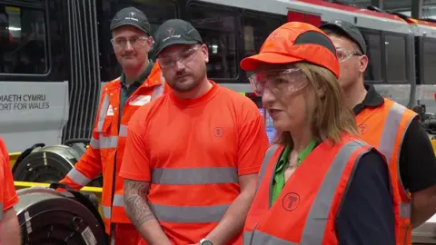 Rachel Reeves wearing orange hi-viz to the right of the photograph, surrounded by workers a train depot to her left. All are wearing protective eye wear.