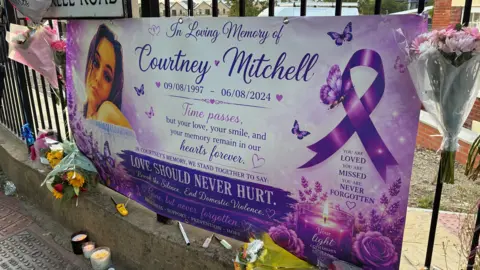 George King/BBC A large purple banner attached to some metal railings. It has Courtney Mitchell written on it and a photo of her. There are some flowers and candles underneath it. 
