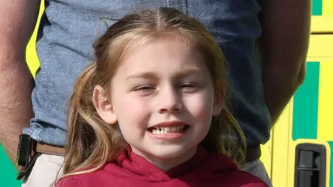 Esmai smiles at the camera. She has brown hair that has been tied back behind her head and she wears a red hoodie. Behind her a man can be seen as well as an ambulance.