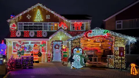 Linda De Santiz Sarum Avenue in Melksham, Wiltshire. A house is covered in Christmas lights. There are large figures including a Santa, a penguin, presents and a dog which are also lit up. 'Ho ho ho' is spelled out in lights, as is 'Merry Christmas' 