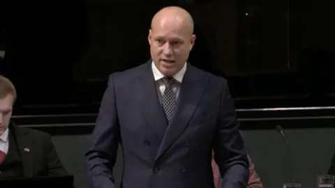 Senedd Cymru Dan Thomas speaking in the Senedd in a double breasted suit and a spotted tie.