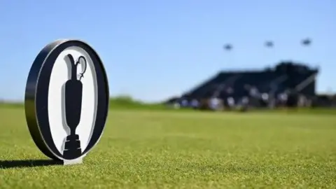 An Open logo coin on the course of Royal Portrush. A blurred bandstand can be seen in the background. 
