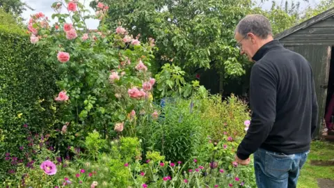 Man wearing glasses, black jumper and blue jeans in his garden overlooking a bed with colourful flowers.