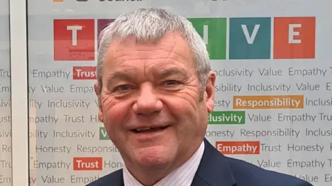 The image shows a person standing in front of a brightly coloured backdrop featuring the branding and values of Herefordshire Council. The individual in the foreground is wearing a dark suit jacket, a light shirt, and a patterned tie. The backdrop appears to be part of a council display wall or promotional board.