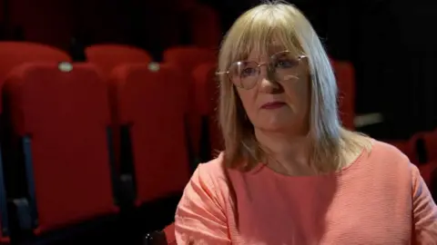 Catriona Cunningham sits in a theatre, wearing a salmon coloured top. She has shoulder length blonde hair and is wearing glasses.