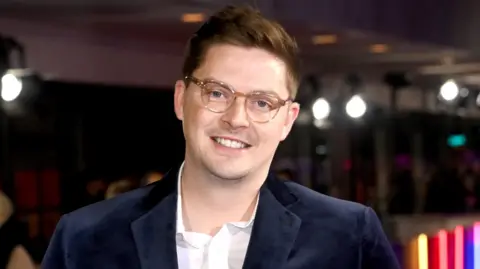 Alex George, who is wearing a blue jacket and glasses