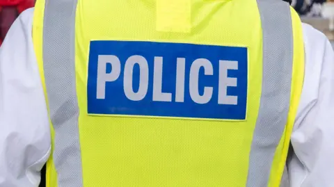A generic shot of a police officer from behind. They are wearing a hi-viz vest with the word "police" printed on it. 