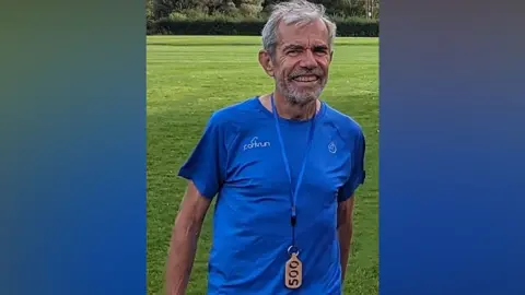 A man in a light blue running top smiles at the camera. He has grey hair and a grey beard. The top has parkrun written across his right chest in white. He is wearing a lanyard around his neck with a wooden pendant on the end, with 500 carved out of it. 