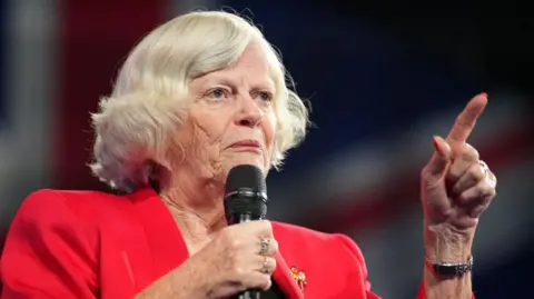 Getty Images Ann Widdecombe wears a red jacket and holds a microphone while speaking at the Reform Party 2024 Conference.