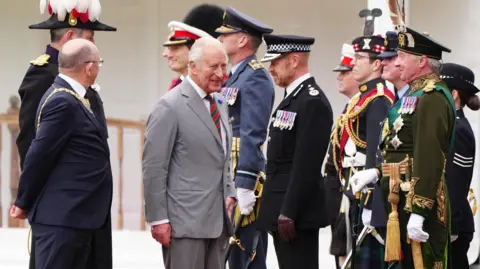 PA Media King Charles III wearing a grey suit and white shirt walks with the Lord Provost of Edinburgh Robert Aldridge, who wears glasses and a dark-coloured suit. The pair are greeted by uniformed figures.