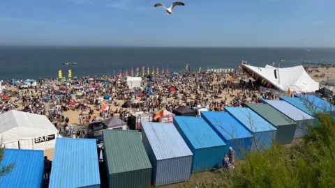 It's a beach front view overlooking the sea. There is a music festival going on, on the beach with crowns and flags with a music stage.