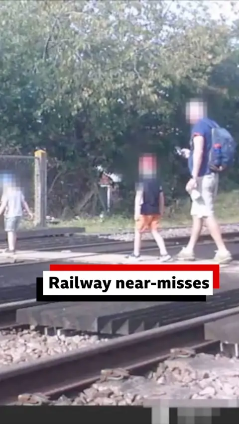 A man on his phone walks across a railway line with two children. The man is looking at his phone while walking across the line at a crossing point. There are two small children in front of him. They are all wearing shorts and T-shirts and their faces have been blurred out.