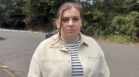 Hannah Irwin stands in front of a road, located next to large bushes and trees, which is cordoned off with police tape. She is looking at the camera with a neutral expression. She has light brown hair, which has been tied back, with a piece at the front covering her face. She is wearing a cream buttoned jacket, with a white and black striped top.