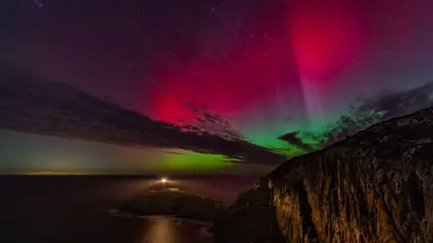 Gail/BBC Weather Watchers A swathe of pink and green lights illuminate Trearddur Bay, dramatically lighting up the cliffs, which stand in the foreground to the right of the image.