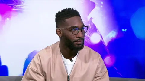 Tinie Tempah has short dark hair and is wearing a beige quarter-zip jumper.