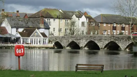 An old stone bridge with arches spans the River Avon at Fordingbridge. A bench and life ring are on the grass-covered near bank. The far bank is clustered with houses.