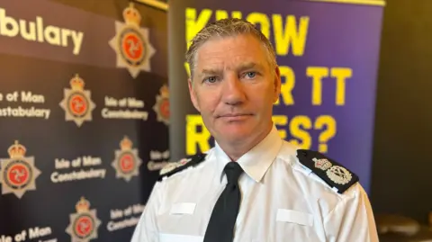 Russ Foster has short grey hair and wears a white shirt, black tie and black lapels. He stands in front of colourful signage which has the Isle of Man Constabulary's emblem.