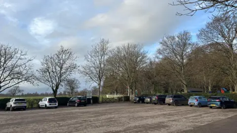 Swindon Borough Council A large outdoor car park in a rural looking setting with a gate and field behind a car park with nine cars parked. There are bare trees lining the car park.