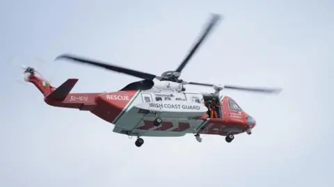 A helicopter flying in the sky. It is red and white in colour and has the words Irish coast guard written in black.