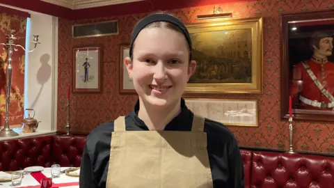 A young woman wearing a black chefs outfit and a khaki apron. She has tied back brown hair and is smiling. In the background, there are red leather lounge seats and a table with a white table cloth that has a red cross on it.