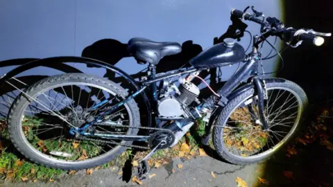 A bicycle is propped up against a wall at night. It is dark in colour and has an engine attached to the cross bar.