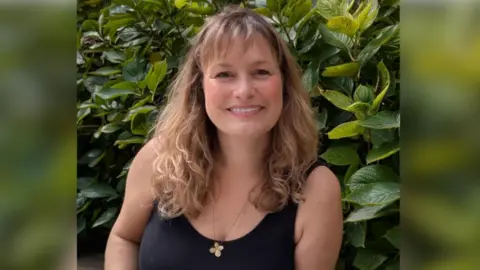 Lisa Timms Lisa Timms, a woman who is sitting outside in front of leafy green foliage. She is looking directly at the camera and smiling and is wearing a black T-shirt and gold necklace.