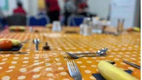 Emma Baugh/BBC A table covered in an orange wipeable tablecloth with white dots. It is laid for meal with knives, forks and spoons, as well as salt and pepper. At each laying is a banana and tomato. People can be glimpsed milling around in the background.