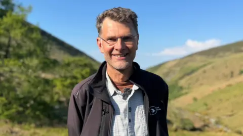 Leigh Morris, who has short grey hair and is wearing glasses. He's wearing a white shirt and black fleece jacket and standing in front of a green valley on a sunny day.