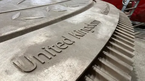 Close-up view of a large circular bronze structure with raised lettering reading 'United Kingdom' along the edge. The surface features engraved leaf patterns. A metal ladder and red panels are visible in the background inside an industrial workshop.