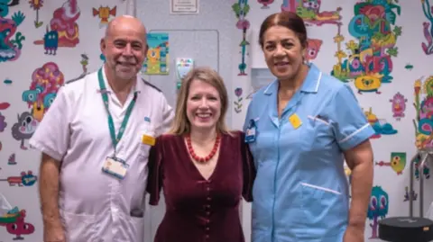 Marie Tidball, who has shoulder length blonde hair, smiles for a photo along with two members of hospital staff. The man to her left, Jonathan Pagdin, is bald with short grey facial hair, while the woman on her right, Joyce Afflick has brown hair pulled back into a bow.