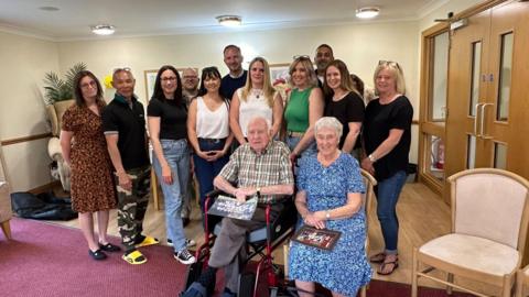 A group of former students standing behind Mr Morgan at the care home and smiling