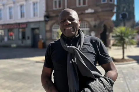 Fitzroy smiling directly into camera, wearing a black t-shirt with a dark grey scarf and a black jacket over his arm. Wrexham high street is visible in the background. 