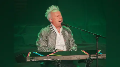 The Cynthia Woods Mitchell Pavilion Howard Jones at a keyboard, performing. He is singing with his eyes closed. He has his trademark spiky blonde hair and a silver sparkled jacket.