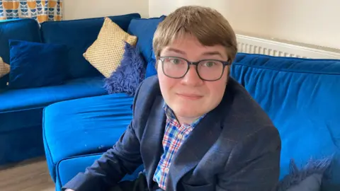 A man with brown hair swept across his forehead is wearing black rimmed glasses and a navy blazer and blue and red shirt. He is sat on a blue corner sofa.
