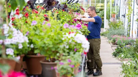 Duncan Mcmillan A gardener in green combat trousers and a blue T-shirt tends to a large display of colourful flowers in a glass house