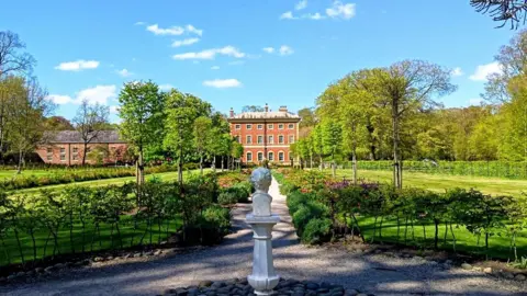 Seaside walker me A three-storey Georgian palladian mansion with high windows stands in the centre background behind a large garden. Trees, shrubs and flowers line a central path with a white stone pillar in the foreground. Some clouds can be seen in the bright blue sky.