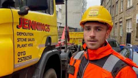 A man in a yellow hard hat and orange reflective jacket standing next to a yellow lorry cab.