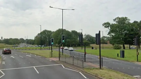 The road junction next to a roundabout. There is a pedestrian crossing with traffic lights.