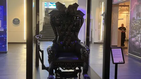 Black ornate chair labeled ‘The Throne of Ozzy Osbourne’ displayed in a glass case inside a museum, featuring bat-wing details and skull armrests, with exhibition panels visible in the background