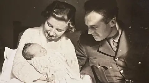 Roseanna Rolph A black and white photograph of a woman cradling a baby and a British soldier, who is looking at the baby.
