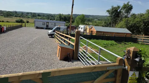 An open area of land with a gravel surface. There is a mobile home on the site along with a caravan and two sheds. In the background are hills and greenery.