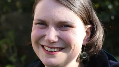 Green Party Sarah Wakefield has shoulder length brown hair and is wearing green earrings.