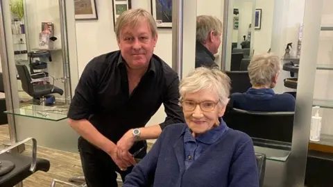 Gillian and Andrew in the salon. Andrew has short grey brown hair and wears a black shirt and trousers. Gillian has short stylish grey hair, and wears glasses and a blue shirt and cardigan.