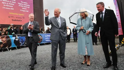 PA Media King Charles smiles and waves. He is wearing a grey suit and pale grey tie. Queen Camilla stands beside him wearing a long blue coat and she carries an umbrella. There are two men in suits either side of them in the photo. Photographers in the background as well as signage for the Fleadh.
