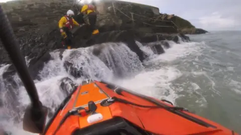 Newquay coastguards The image shows a rescue scene on a rocky shoreline. An orange lifeboat is positioned in choppy water close to the rocks, with waves breaking against the stone steps and ledges. Two rescuers in yellow and red gear with helmets are standing on the rocks above the waterline, appearing to assist with an operation. A stone wall and safety railings run along the top of the slope.