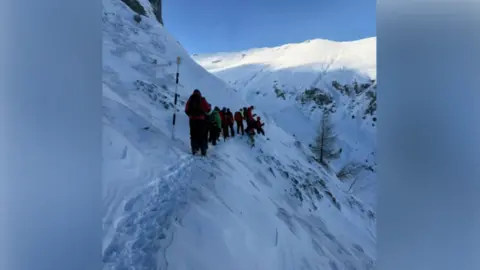 Salvamont Brașov A line of people follow a mountain path in snow. Mountains can be seen all around with rescuers in winter gear.