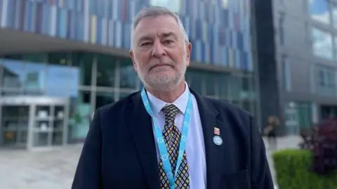 BBC A man with short grey hair ,wearing a navy blazer, pale blue shirt and black, blue and yellow tie is standing in front of the Doncaster council Waterdale building.